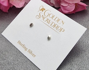 May include: A pair of sterling silver stud earrings with a smooth, oval design. The earrings are displayed on a white card with the text "Golden Snowdrop" and "Sterling Silver" in gold lettering. Pink flowers are in the background.