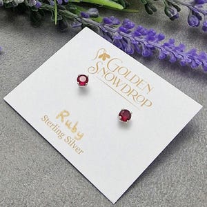 May include: Ruby stud earrings on a white card with the text "Golden Snowdrop" and "Ruby Sterling Silver". The earrings feature a silver setting and a deep red gemstone. The background includes purple flowers.