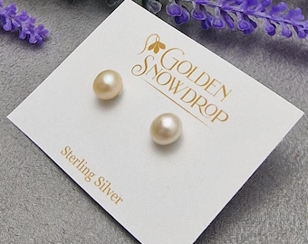 Peut inclure: Une paire de boucles d'oreilles en perles de couleur pêche clair sur une carte blanche avec le texte "Golden Snowdrop" et "Sterling Silver". Les boucles d'oreilles sont rondes et reposent sur une surface grise avec des fleurs violettes.