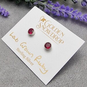 Puede incluir: Pendientes de plata de ley con gemas de rubí cultivadas en laboratorio. Los pendientes se muestran en una tarjeta blanca con texto dorado que dice "Golden Snowdrop", "Lab Grown Ruby" y "Sterling Silver". Flores moradas en el fondo.