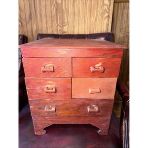 May include: A small wooden chest of drawers featuring a distressed red finish with visible wood grain. The unit has five drawers arranged in three rows, each equipped with a simple wooden handle. The base has two arched cutouts at the bottom.