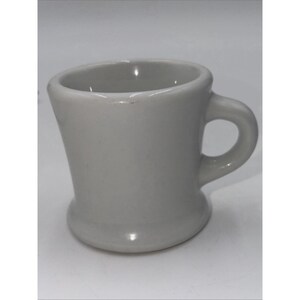 May include: A gray ceramic mug with a curved body and a rounded handle. The mug has a wide opening and a flared base. The mug is a solid, neutral color, suitable for coffee or tea.