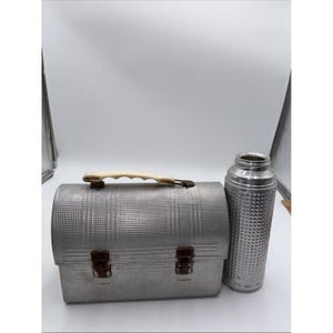 May include: A rectangular metal lunch box with a textured grid pattern, a cream-colored plastic handle, and two brown metal latches. Beside the lunch box stands a matching cylindrical metal thermos with a textured grid pattern on its exterior surface.