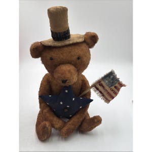 Könnte beinhalten: Ein brauner Plüschteddybär mit einem beigen Zylinder, der ein dunkles Streifenband hat. Der Bär hält einen dunkelblauen Stoffstern mit weißen Sternen und eine kleine, verwitterte amerikanische Flagge an einem Holzstab. Schwarze Knopfaugen.