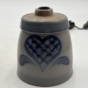 Pode incluir: Uma base de candeeiro cilíndrica em grés com acabamento cinzento claro e um design de coração pintado à mão em azul escuro com um padrão de grelha. A parte superior é de um cinzento ligeiramente mais escuro com uma abertura central.