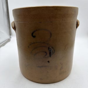 Pode incluir: Um pote cilíndrico de grés na cor castanha com duas alças laterais e um número três pintado à mão em azul escuro na frente. O recipiente tem um acabamento fosco suave com variações sutis de cor e uma borda superior ligeiramente elevada.