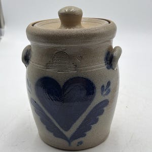 Puede incluir: Una vasija de gres beige con tapa y dos asas laterales. El frente presenta un gran diseño de corazón pintado en azul oscuro con detalles de hojas. Una marca estampada dice ROWE POTTERY WORKS CAMBRIDGE WIS 1986, con una pequeña marca en forma de V.