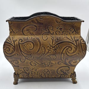 May include: A rectangular metal planter with a curved top edge and four decorative feet. The exterior features a raised, embossed floral and scroll pattern in a dark brown finish over a muted gold base, creating a textured, aged appearance.