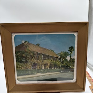 May include: A framed vintage color photograph of a historic building covered in climbing vines, with the text "Oldest House in the United States St. Augustine" printed at the bottom. The wooden frame has a light brown finish and a wide border.