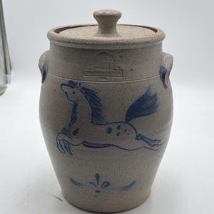 May include: A beige stoneware crock with a lid and two side handles. The exterior features a blue hand-painted horse illustration and a small decorative flourish below. A stamp near the top rim reads "ROWE POTTERY WORKS CAMBRIDGE WIS 1984".