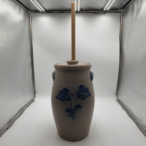 Puede incluir: Una mantequera cilíndrica de gres con un acabado en color tostado claro y moteado. Presenta un motivo floral pintado a mano en azul oscuro en la parte frontal, dos asas laterales, una tapa a juego y un largo mango de madera vertical.