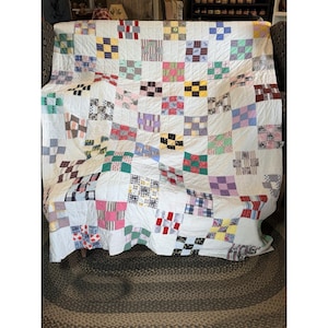 May include: A patchwork quilt featuring a grid of white fabric squares alternating with various multi-colored patterned fabric squares. The quilt displays a variety of colors including red, blue, yellow, green, pink, and purple in plaid, floral, and striped prints.
