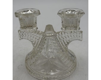 May include: A clear pressed glass double candlestick holder featuring a central pointed finial and two candle cups. The base and arms are decorated with a geometric diamond and star pattern, creating a textured, multifaceted surface.