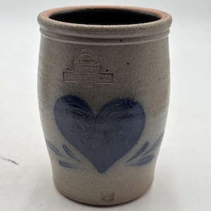 Puede incluir: Una vasija de gres cilíndrica con un acabado de color tostado claro y moteado. La parte frontal presenta un diseño de corazón pintado en azul oscuro con pequeños detalles de hojas. Un sello impreso dice: HANDMADE, ROWE POTTERY WORKS, CAMBRIDGE WIS.