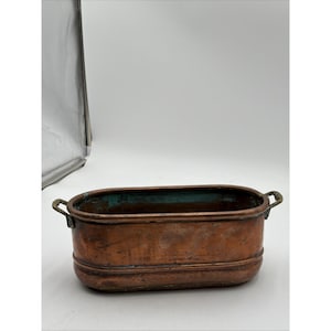 May include: An oval copper planter with two small metal handles on the sides. The exterior has a weathered, warm metallic finish with visible surface texture. The interior shows signs of oxidation with a distinct teal green patina throughout.
