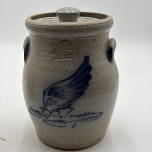 May include: A beige stoneware crock with a lid, featuring two side handles and a blue cobalt bird illustration. The front displays a stamped maker's mark that reads "ROWE POTTERY WORKS CAMBRIDGE WIS 1986". The vessel has a speckled texture.