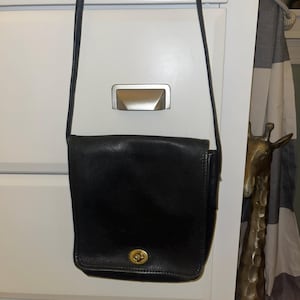 May include: A black leather crossbody bag with a rectangular flap, a thin shoulder strap, and a gold-toned metal turn-lock closure on the front. The bag features a smooth texture and a structured design with visible stitching along the edges.