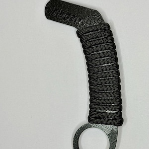 May include: A black karambit-style training tool featuring a textured handle wrapped in black paracord. The handle includes a finger ring at the base and a curved top section with the embossed text "FIDGET" visible on the surface of the black material.