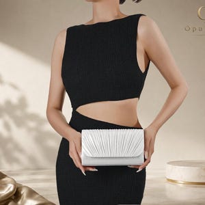 May include: A white rectangular clutch bag featuring a pleated flap design with a smooth, satin-like finish. The clutch is held horizontally and displays a structured, folded texture across the front panel, providing a clean and minimalist aesthetic.