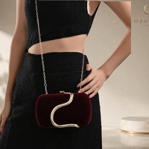 Könnte beinhalten: Eine tiefburgunderrote Samt-Clutch mit einem goldfarbenen, schlangenförmigen Verschluss und einem dünnen Schulterriemen aus Goldkette. Die rechteckige Tasche hat abgerundete Kanten und eine weiche Textur, akzentuiert durch das metallische Schlangendetail.