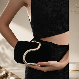 Puede incluir: Un bolso de mano tipo clutch de terciopelo negro con un asa de metal dorado en forma de serpiente que se curva sobre la superficie frontal. El bolso tiene una forma rectangular redondeada y lisa. El logo OpulAura es visible en la esquina superior.
