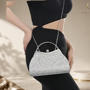 Può includere: Una borsa a mano argentata glitterata con struttura rigida, manico superiore arrotondato in metallo argentato e chiusura decorativa con cristalli incastonati. La borsa include una sottile tracolla a catena argentata e il logo OpulAura.