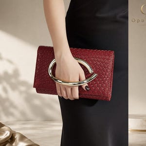 Pode incluir: Uma bolsa clutch retangular vermelho-escura com textura trançada e uma grande alça de metal dourado liso em forma de oval alongada. A bolsa é segurada horizontalmente, exibindo seu design estruturado e o logotipo OpulAura no canto.