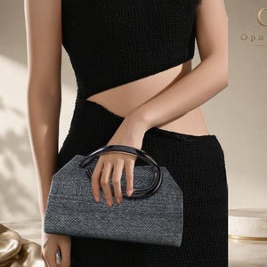 Puede incluir: Un bolso de mano gris oscuro estructurado con una superficie texturizada y un asa negra lisa y redondeada. El bolso presenta una forma geométrica trapezoidal y lo sostiene una persona que viste un vestido negro sin mangas con un corte lateral.