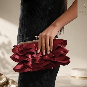 May include: A deep burgundy clutch bag featuring multiple layers of ruffled fabric and a gold-toned metal frame with a ball clasp closure. The brand name OpulAura is visible in the top right corner of the image.