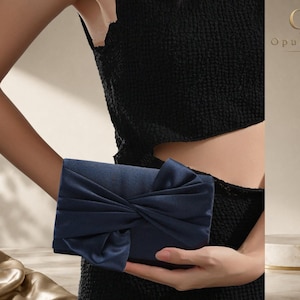 Può includere: Una pochette in raso blu navy con un design a nodo plisato sulla patta anteriore. La borsa da sera rettangolare presenta una finitura liscia e lucente e una forma strutturata, con il marchio OpulAura visibile nell'angolo in alto a destra.