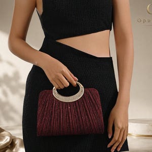 May include: A deep burgundy clutch bag with a pleated, shimmering texture and a semi-circular metallic handle encrusted with clear crystals. The bag features a structured, rectangular silhouette and is held in front of a black textured dress.