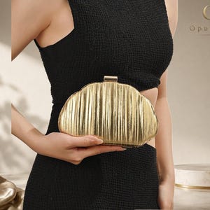 Peut inclure: Une pochette dorée en forme de coquillage avec une texture métallique à côtes verticales et un fermoir rectangulaire sur le dessus. L'accessoire présente une silhouette arrondie et incurvée ainsi qu'une finition polie et réfléchissante.