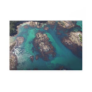 May include: An aerial view of a coastal landscape featuring turquoise water, rocky islands, and patches of brown seaweed. The image captures a serene scene with a forest in the background, showcasing natural beauty.