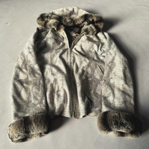 Könnte beinhalten: Eine Jacke mit Reißverschluss und einem cremefarbenen sowie braunen Schlangenledermuster auf dem gesamten Körper. Das Kleidungsstück verfügt über eine Kapuze und Ärmelbündchen mit dickem, strukturiertem braunem Kunstfellbesatz.