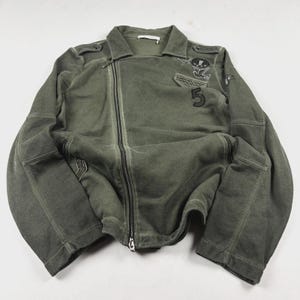 Peut inclure: Une veste de style moto vert olive avec une fermeture éclair frontale asymétrique, un col rabattu et des manches longues. La veste est ornée d'écussons brodés, dont un oiseau et le chiffre 5, ainsi que de clous métalliques décoratifs.