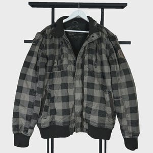 May include: A gray and black checkered jacket featuring a full-length front zipper, a high collar, and ribbed cuffs and hem. The jacket includes two vertical zippered chest pockets, two lower side pockets, and a small patch on the left sleeve.