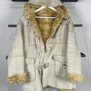 May include: A beige corduroy hooded jacket featuring a tan faux fur lining, cuffs, and hood trim. The jacket includes a front closure with multiple adjustable buckle straps and two large patch pockets on the front with buckle flap closures.