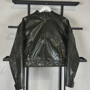 May include: A dark olive green jacket featuring a snakeskin texture pattern throughout. The jacket has a stand-up collar, a full-length front zip, and two zipped pockets on the front. It is displayed on a white hanger against a black frame.