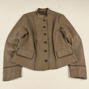 May include: A taupe-colored jacket featuring a mandarin collar, a front button closure with six dark buttons, and two front flap pockets. The garment is accented with decorative stitching along the seams and dark brown trim on the cuffs and pockets.