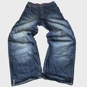May include: Dark blue denim jeans with a wide-leg silhouette and a faded wash effect. The jeans feature a button fly, belt loops, and a small rectangular label on the lower left leg with the text "rocca-wear" in white lettering.