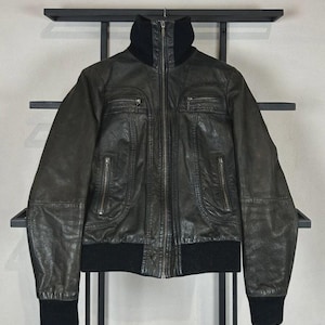 May include: A black leather jacket featuring a high ribbed collar, ribbed cuffs, and a ribbed hem. The jacket includes a central metal zipper closure, two zippered chest pockets, and two additional zippered pockets located at the waist area.
