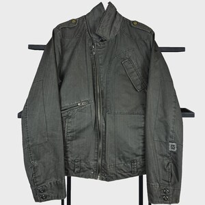 May include: A dark charcoal gray jacket featuring an asymmetrical front zipper closure, a high collar, and shoulder epaulets. The garment includes a zippered chest pocket, a flap pocket, and a small square patch with the number 65 on the left sleeve.