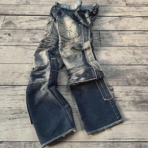 May include: Blue denim jeans featuring a patchwork design with multiple rectangular panels of varying shades of blue. The jeans have a distressed finish, visible stitching, and frayed raw edges at the bottom hems of the pant legs.