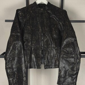 Puede incluir: Una chaqueta negra con un patrón texturizado de piel de serpiente en oro metálico y negro. La chaqueta presenta un cuello alto, mangas largas y un cierre de cremallera central en la parte delantera. La prenda tiene una costura en la cintura.
