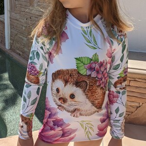 May include: White long-sleeved shirt with a watercolor print of hedgehogs, flowers, and leaves. The shirt has a crew neck and long sleeves.