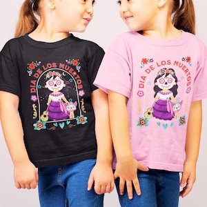 May include: Black and pink t-shirts with a Dia de los Muertos graphic. The design features a girl with a sugar skull face, a purple dress, and a guitar, surrounded by floral elements and the text "Dia de los Muertos".