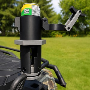 Peut inclure: Un porte-boisson en métal noir et gris monté sur un support de véhicule, doté d'un bras latéral réglable et d'une base cylindrique. Une canette de boisson jaune et verte est insérée dans le support, affichant un texte de valeurs nutritionnelles.