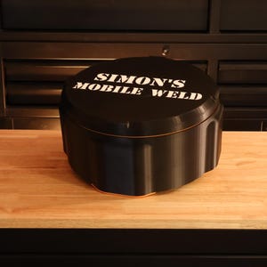 May include: Black, rounded container with the text "SIMON'S MOBILE WELD" in white. The container sits on a wooden surface, with a dark background.