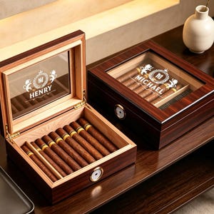 May include: Two dark wood humidors with glass tops and gold-tone hinges. One humidor is open, revealing a cedar interior filled with brown cigars. The other is closed, displaying the name MICHAEL and a crest. Both feature a round analog hygrometer.