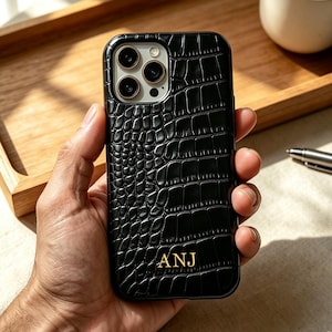 Puede incluir: Una funda de smartphone negra con un patrón texturizado de piel de cocodrilo y las letras doradas ANJ grabadas en la parte inferior. La funda cuenta con un recorte para un sistema de cámara de triple lente en la parte posterior.